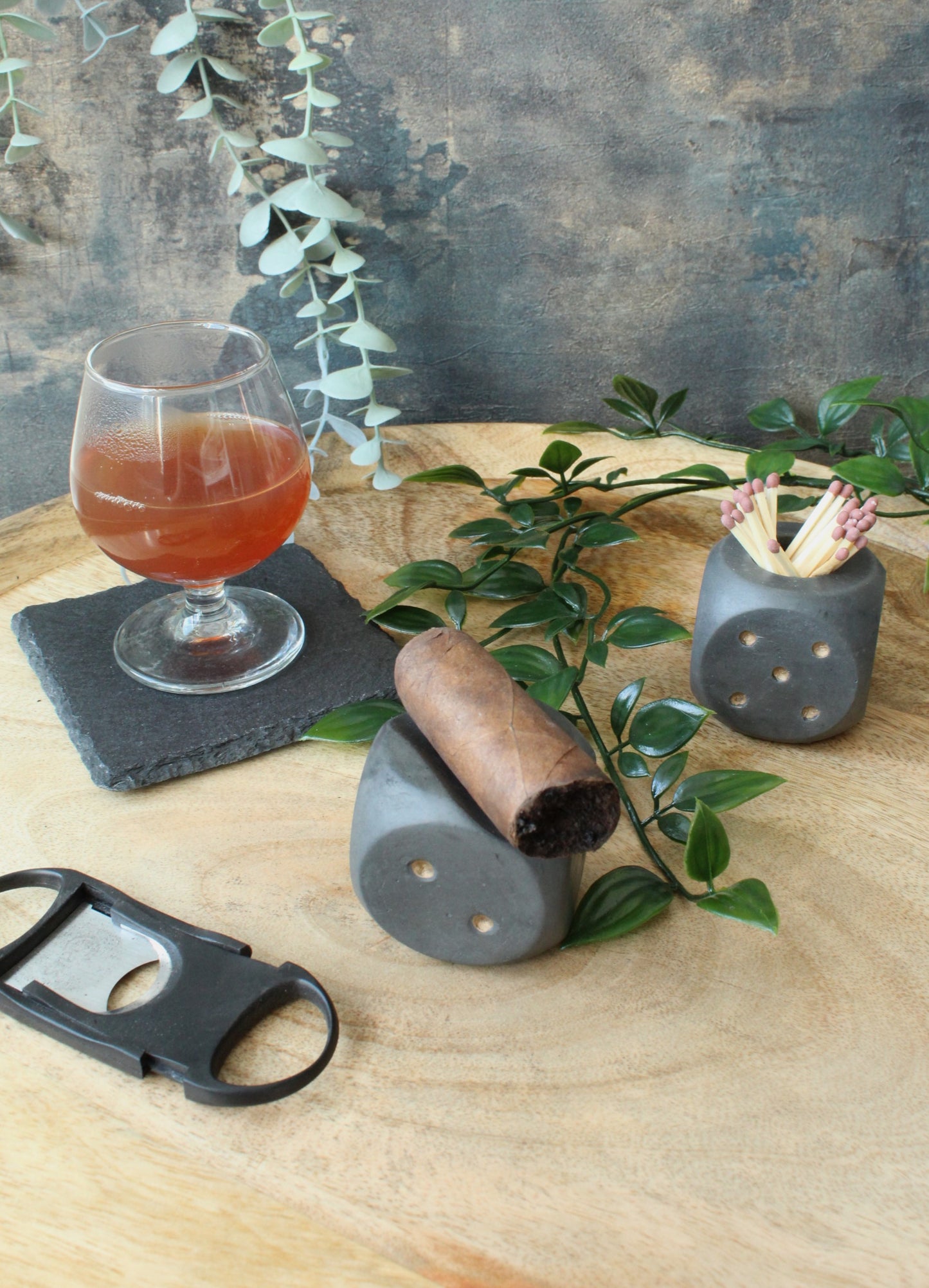 A cosy table-top scene featuring a glass of whiskey on a black slate coaster, a dark cigar resting on a dice-shaped holder, and another dice-shaped container filled with matches. A cigar cutter lies nearby on the wooden surface, surrounded by green leaves and a rustic, textured backdrop with hanging eucalyptus.