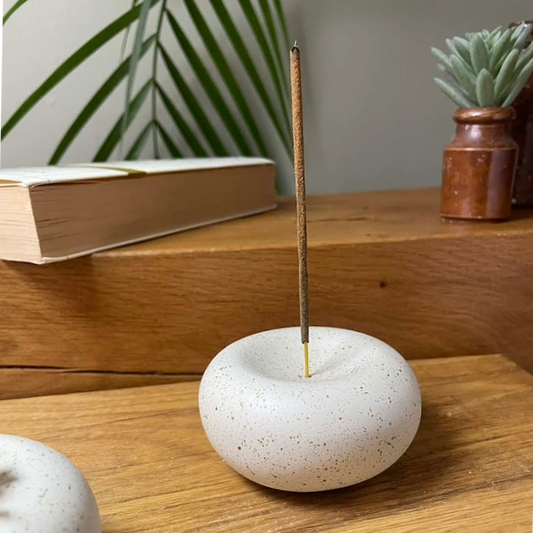 White speckled stone incense holder with a single stick on a wooden surface, with a book and plant in the background.