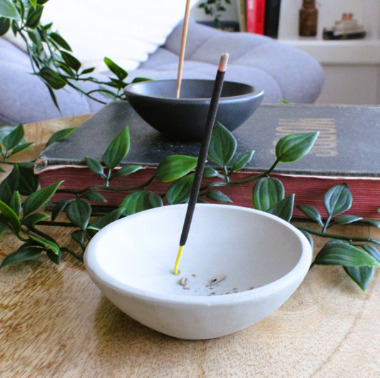 Simple, minimalist natural stone incense stick holder in the shape of a dish. In the background is a similar dish sitting on an old book finished in matte black. Some green leaves gently surround both dishes