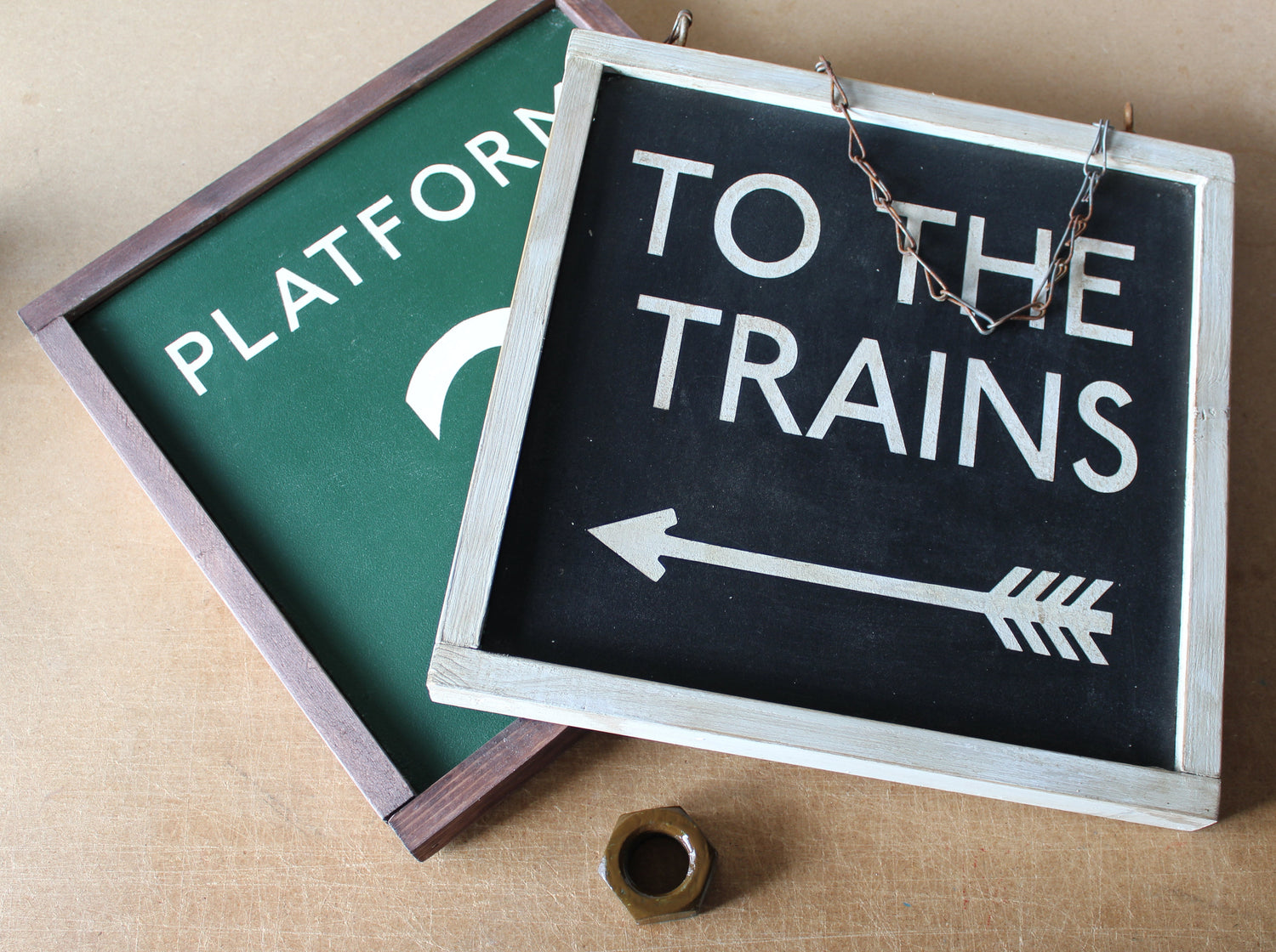 A rustic framed chalkboard sign reading 'TO THE TRAINS' with a white arrow, shown on a tan surface with another green "platform" sign partially visible behind it.