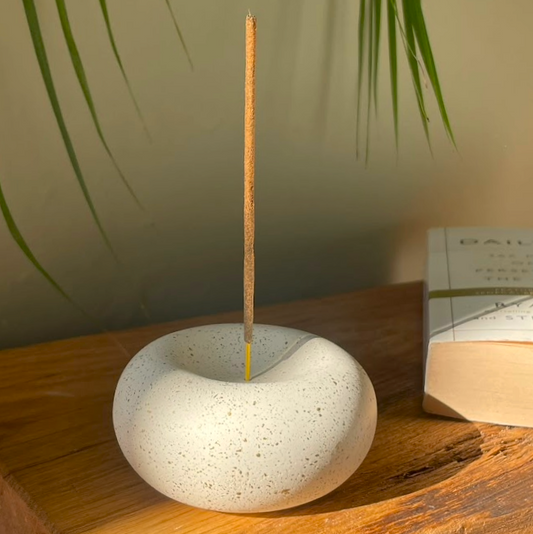 Stone incense holder with a single incense stick on a wooden surface with a plant in the background.