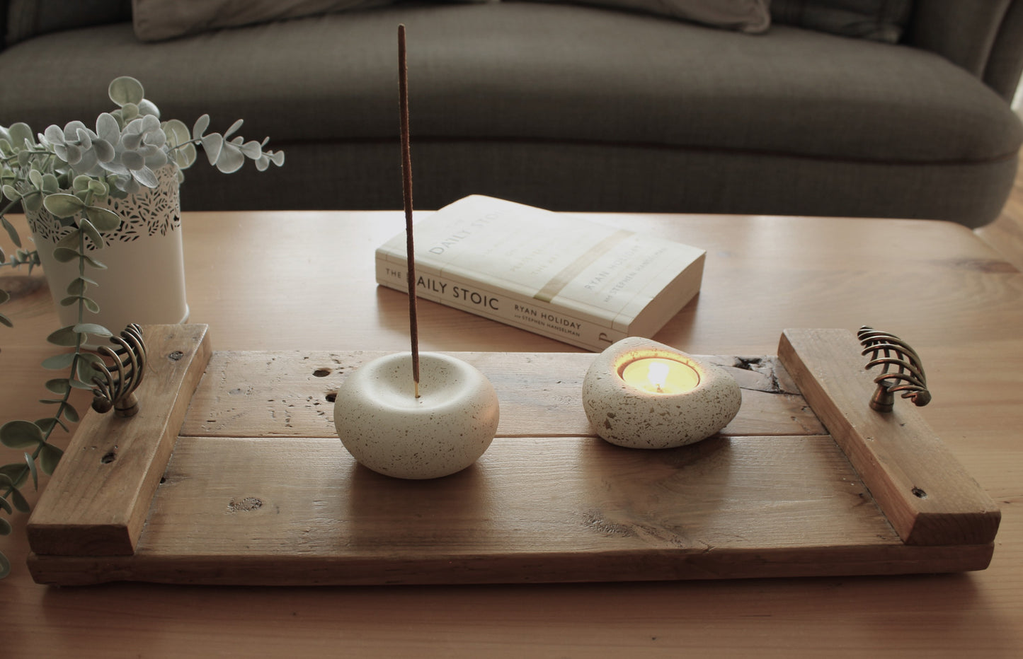 White stone incense holder with a candle on a wooden tray, with a book and plant in the background.