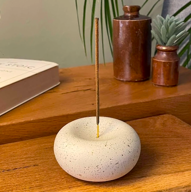 Stone incense holder with a single stick on a wooden surface with books and jars in the background.
