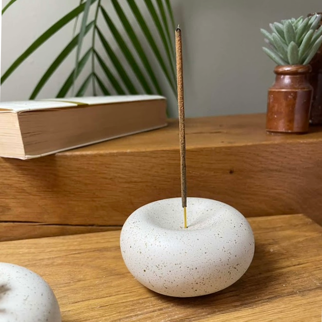 White speckled stone incense holder with a single stick on a wooden surface, with a book and plant in the background.