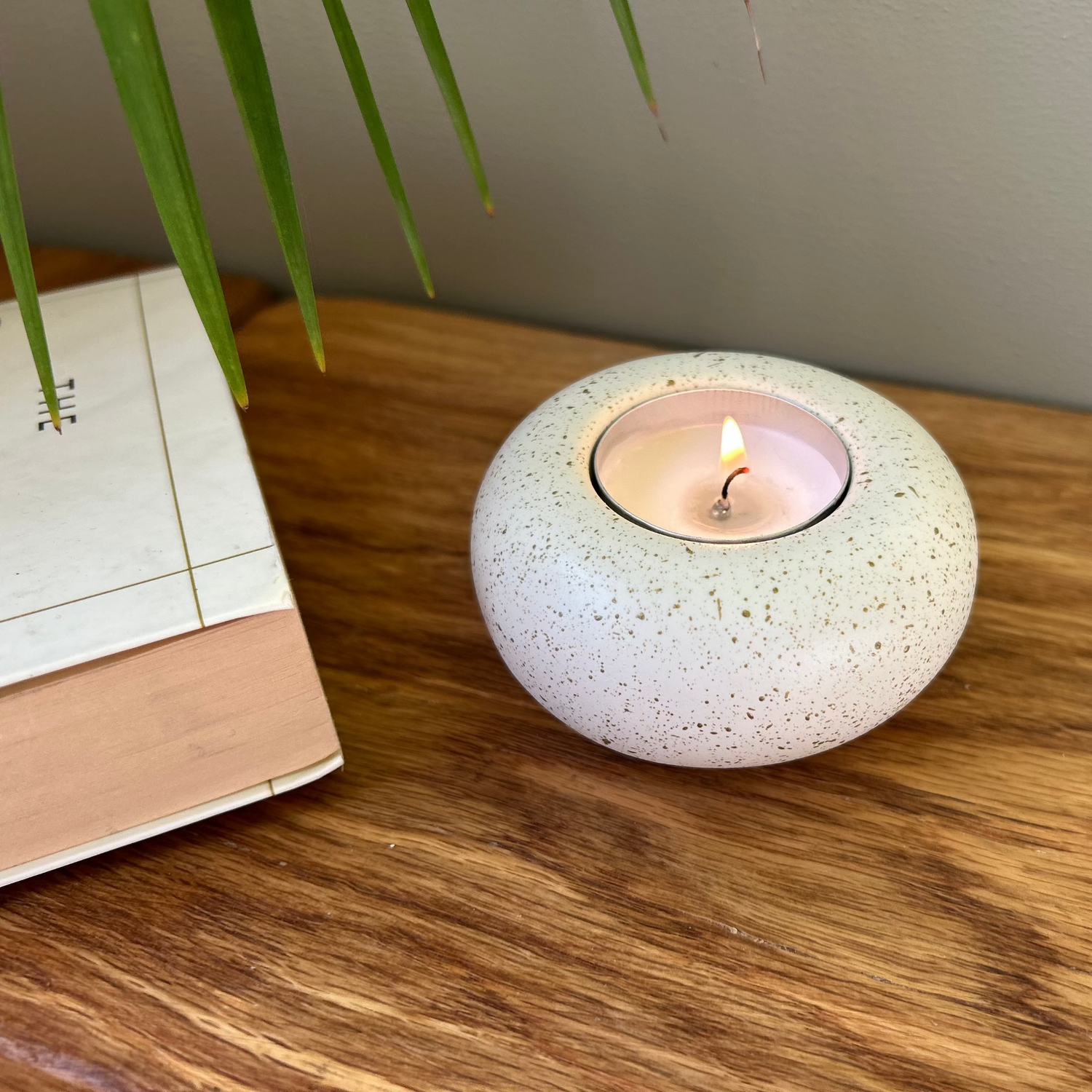 A round speckled ceramic candle holder containing a lit tealight candle, placed on a wooden tabletop next to a book and green plant leaves.
