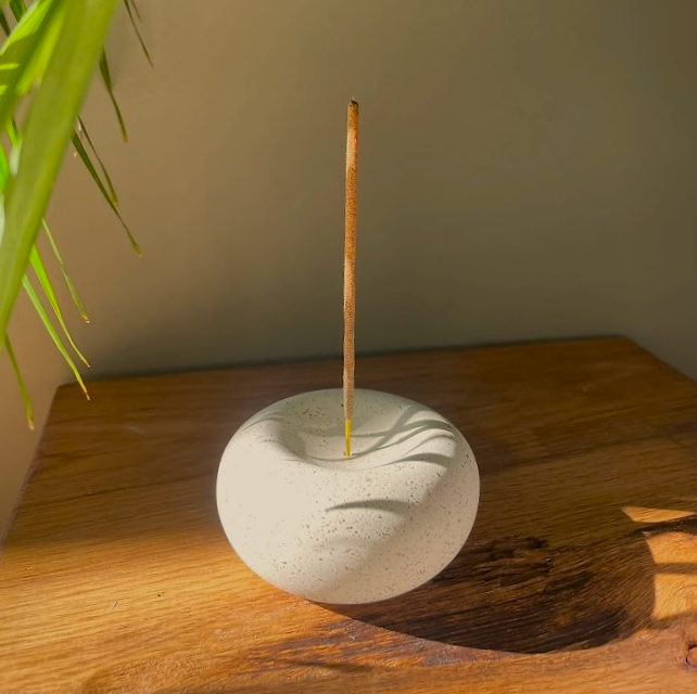 Stone incense holder with a single incense stick on a wooden surface, with a plant in the corner.