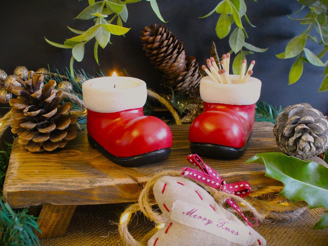 Pair of red and white cast stone Santa boots, one holding matches, one holding a lit tea light