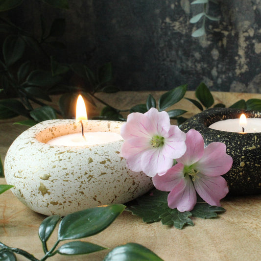 A pair of handmade candle holders, one white with gold specks and the other black with gold specks, each containing a lit candle. They are placed on a wooden surface, surrounded by green leaves and pink flowers. The candles are gently glowing, creating a cozy and natural atmosphere.
